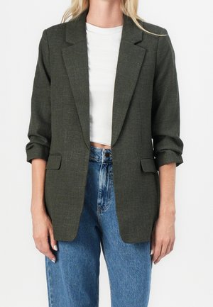 Kobieta ubrana w ciemnozieloną marynarkę na białym krótkim topie i niebieskie, wysokie dżinsy, z podwiniętymi rękawami marynarki.