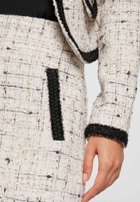 Blazer de tweed con un patrón texturizado en blanco y negro, que presenta un acabado negro en los puños y los bolsillos. El primer plano muestra el detalle de la tela.
