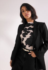 Veste noire sur mesure portée sur un haut noir et beige à motifs, associée à un jean noir taille haute et une fine ceinture dorée. Textures lisses dans l'ensemble.