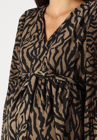 Žena na sobě má šaty se zebrovaným vzorem v hnědé a černé barvě, s výstřihem do V a zavázaným opaskem v pase, opatřené plisovanými dlouhými rukávy.