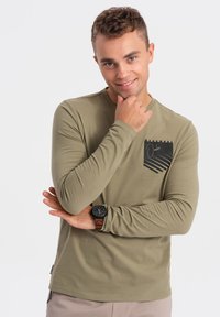Langarm-Olivgrünes T-Shirt aus Baumwolle mit einem schwarzen geometrischen Brusttaschendesign. Verfügt über einen runden Halsausschnitt und taillierte Ärmel.