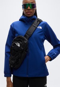 Veste imperméable bleue avec capuche, dotée d'une fermeture éclair à l'avant et de poignets élastiques. Sac bandoulière noir avec logo. Pantalon foncé, lunettes de soleil oversize.