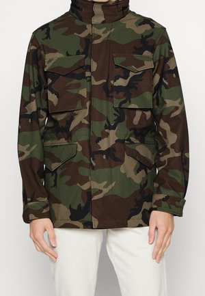 Osoba nosząca zielono-brązową kurtkę kamuflażową z czterema przednimi kieszeniami z klapami oraz jasne spodnie.