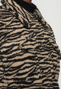 Beige knapparjacka med svarta tigerränder. Har en krage och två framfickor med rundade knappar. Mjuk, strukturerad yta.