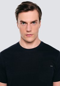 Młody mężczyzna o krótkich brązowych włosach, ubrany w czarny t-shirt z okrągłym dekoltem, patrzący przed siebie z neutralnym wyrazem twarzy.