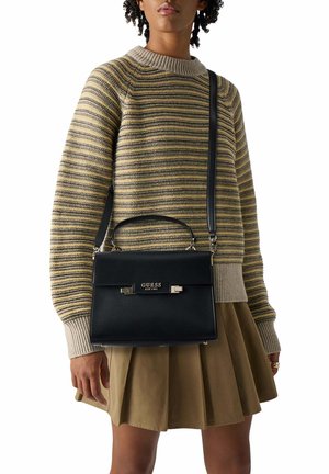 Personne portant un pull rayé beige et gris et une jupe plissée beige, portant un sac à main Guess noir avec une poignée supérieure et une bandoulière.