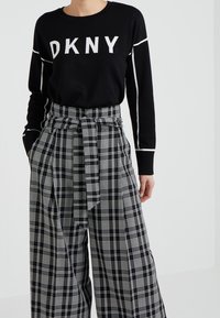 Schwarzer Pullover mit weißem "DKNY"-Logo, kombiniert mit weit geschnittenen Schwarz-Weiß-karierten Hosen mit gebundenem Bund und weichem Stoff.