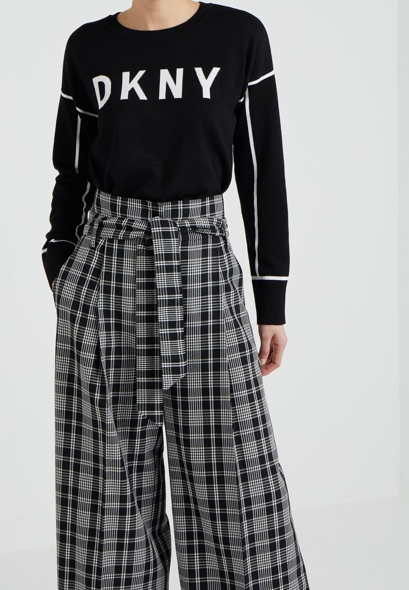 Schwarzer Pullover mit weißem "DKNY"-Logo, kombiniert mit weit geschnittenen Schwarz-Weiß-karierten Hosen mit gebundenem Bund und weichem Stoff.