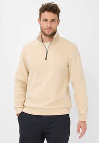 Uomo con i capelli ricci corti, che indossa un pullover beige con zip fino al quarto e pantaloni neri, in piedi con una mano in tasca su uno sfondo bianco.