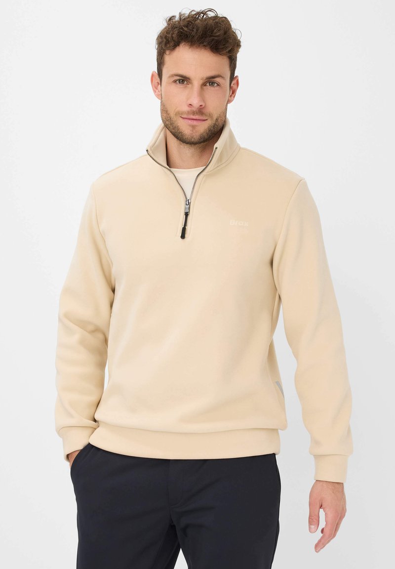 Uomo con i capelli ricci corti, che indossa un pullover beige con zip fino al quarto e pantaloni neri, in piedi con una mano in tasca su uno sfondo bianco.