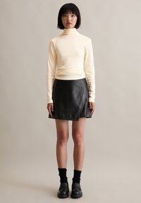 Hellbeige Rollkragenpullover mit schlanker Passform, kombiniert mit einem schwarzen Leder-Wickelrock, ergänzt durch schwarze klobige Schuhe und gemusterte Socken.