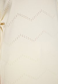 Pulover din tricot crem, cu modele în zigzag formate din mici găuri și accente subtile de maro, având un finisaj moale și texturat.