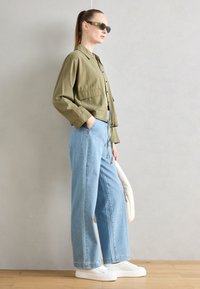 Chemise verte olive à manches longues avec ceinture nouée, associée à un jean bleu clair à jambes larges et des baskets blanches. Accessoirisée avec des lunettes de soleil.