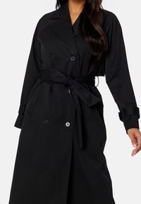 Černý trenčkot s dvojitým zapínáním na knoflíky, páskem v pase, knoflíkovými detaily a dlouhými rukávy s manžetami. Hladká textura látky.