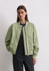 Light green bomber jacket with a smooth texture, ribbed cuffs, and a zippered pocket on the left sleeve. Paired with a black top and pants.