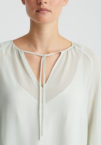 Hellgrüne, durchsichtige Bluse mit V-Ausschnitt und Halsband. Weicher Stoff mit fließender Textur und subtilem Glanz. Verfügt über zarte Falten entlang des Ausschnitts.