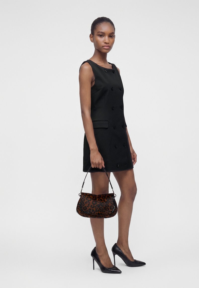 Femme portant une robe noire sans manches boutonnée, tenant un sac à main imprimé léopard et portant des talins hauts noirs sur un fond uni.