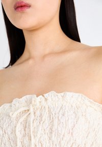 Top senza spalline di colore crema con tessuto testurizzato, caratterizzato da un colletto a volant e un dettaglio con coulisse nella parte anteriore. Materiale liscio e leggero.
