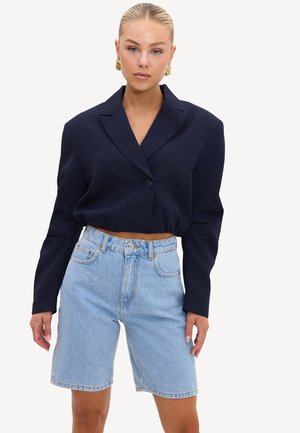 Femme aux cheveux plaqués en arrière, portant des boucles d'oreilles en or, un blazer cropped bleu marine et un short en denim bleu clair jusqu'aux genoux.