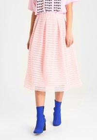 Persona con una falda midi de rayas rosas, camiseta rosa con el texto "boys" y botines azules brillantes con tacón caminando sobre un fondo blanco.