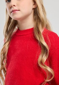 Red knitted sweater with a ribbed neckline. The texture appears soft, with long, wavy, blonde hair partially visible beside the garment.