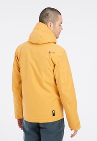 Geel waterdichte jas met capuchon, voorzien van een logo op de achterkant en verstelbare manchetten. Draag het over grijze broek, tegen een witte achtergrond.