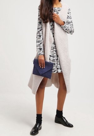 Světlý šedý vlněný svetr s otevřeným předním dílem, ve spojení s vzorovanými šaty. Drží námořnickou kabelku, stylizováno s černými ponožkami a lesklými botami.