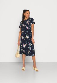 Vestuário floral azul-marinho com mangas curtas, apresentando uma cintura ajustada e uma saia fluida. Combinado com saltos abertos amarelos e uma bolsa pequena.