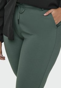 Grønne, elastiske joggebukser med sidelommer, snor i midjen og en glatt tekstur, vist sammen med en svart bluse.