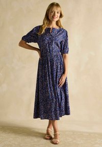 Modré květinové maxi šaty s krátkými nabíranými rukávy, priliehavým pásom a detailom s gombíkmi. Hladká látka s zložitými červenými a bielými vzormi. Béžové sandále.