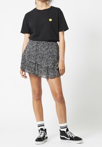 Mustat T-paita, jossa on keltainen hymyilevä naama, yhdistettynä musta-valkoiseen kukalliseen volangihameeseen. Mallilla on päällä mustat tennarit, joissa on valkoiset pohjat ja raidalliset sukat.