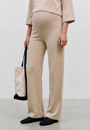 Persona che indossa pantaloni larghi beige e top, tiene in mano una borsa tote beige e nera, in piedi su pavimento grigio con scarpe slip-on nere.
