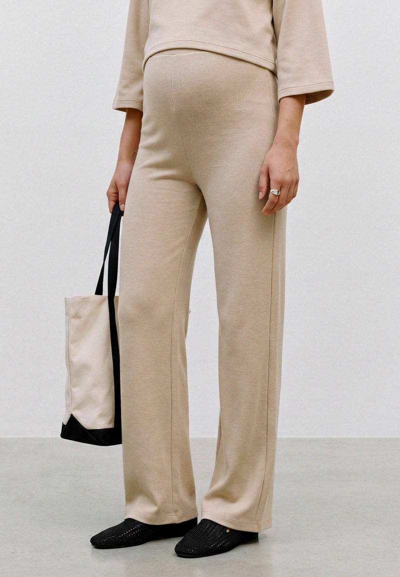 Personne portant un pantalon ample beige et un haut, tenant un sac fourre-tout beige et noir, debout sur un sol gris avec des chaussures noires sans lacets.