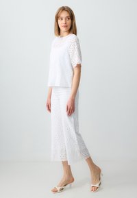 Witte eyelet-top met korte mouwen, gecombineerd met bijpassende brede eyelet-broek. De outfit beschikt over ingewikkelde uitsnijdingen en gegolfde randen.