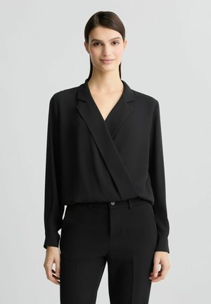 Blouse noire à enroulement en tissu lisse, dotée d'un col et de manches longues, portée avec un pantalon noir taille haute. Design simple et élégant.