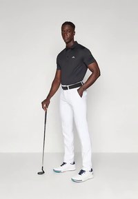 Μαύρο πόλο πουκάμισο με μικρό λογότυπο, άσπρο παντελόνι και λευκά παπούτσια γκολφ με μπλε λεπτομέρειες. Κρατάει ένα γκολφ κλαμπ σε όρθια στάση.