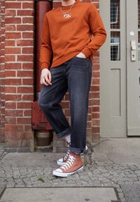 Sudadera de color naranja óxido con un pequeño gráfico, vaqueros oscuros de mezclilla con puños remangados y zapatillas altas marrones con detalles en blanco.