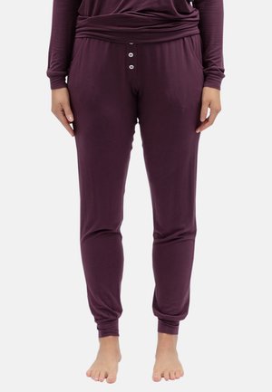 Personne portant un haut à manches longues violet foncé et un pantalon de jogging assorti avec trois boutons et des poches, debout pieds nus sur un fond uni.