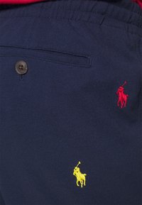 Pantalones cortos de tela azul marino con cierre de botón, que presentan logotipos de polo bordados en rojo y amarillo en la parte frontal. Material texturizado con un ajuste relajado.