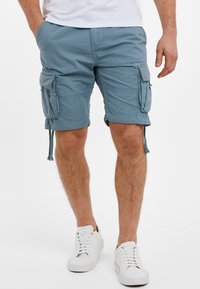 Muž v modrých cargo šortkách a bílých teniskách stojí s rukama částečně v kapsách na bílém pozadí.