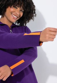Vrouw die glimlacht, gekleed in een paarse lange mouwen top met oranje strepen op de mouwen, die een arm naar voren steekt tegen een witte achtergrond.