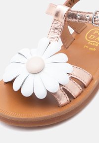Καφέ σανδάλι παιδικό με μεταλλικές ροζ χρυσές λουρίδες και μεγάλη λευκή μαργαρίτα ως διακόσμηση στην περιοχή των δαχτύλων.
