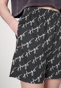 Shorts noirs avec motif logo "Hugo" blanc, taille élastique, poches latérales et texture douce. Design fait main, décontracté pour le confort.