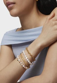 Pulseras de oro y perlas apiladas en una muñeca, mostrando diferentes tamaños, texturas y acabados sobre una tela de color azul claro. Diseño minimalista.