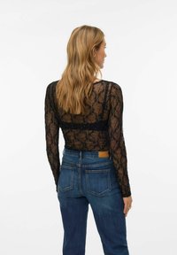 Kvinne med langt blondt hår iført en gjennomsiktig svart blonder overdel og blå denimjeans, sett bakfra mot en ensfarget hvit bakgrunn.