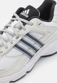 Scarpa sportiva con una tomaia in mesh grigio chiaro, accenti in suede grigio, tre strisce grigio scuro e un collo imbottito nero con il logo Adidas.