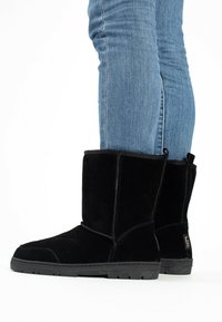 Stivaletti neri in suede con suola piatta in gomma, caratterizzati da un design semplice e un pratico inserto laterale, esposti accanto a un paio di jeans blu.