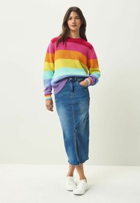 Pull en maille avec des rayures horizontales arc-en-ciel dans des teintes de rose, orange, jaune, vert et bleu ; associé à une jupe maxi en denim et des baskets blanches.