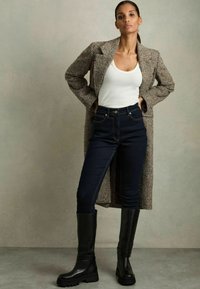 Brun rutigt långkappa, vit linne, mörkblå jeans och knähöga svarta stövlar med kraftiga sulor. Texturen varierar mellan materialen.