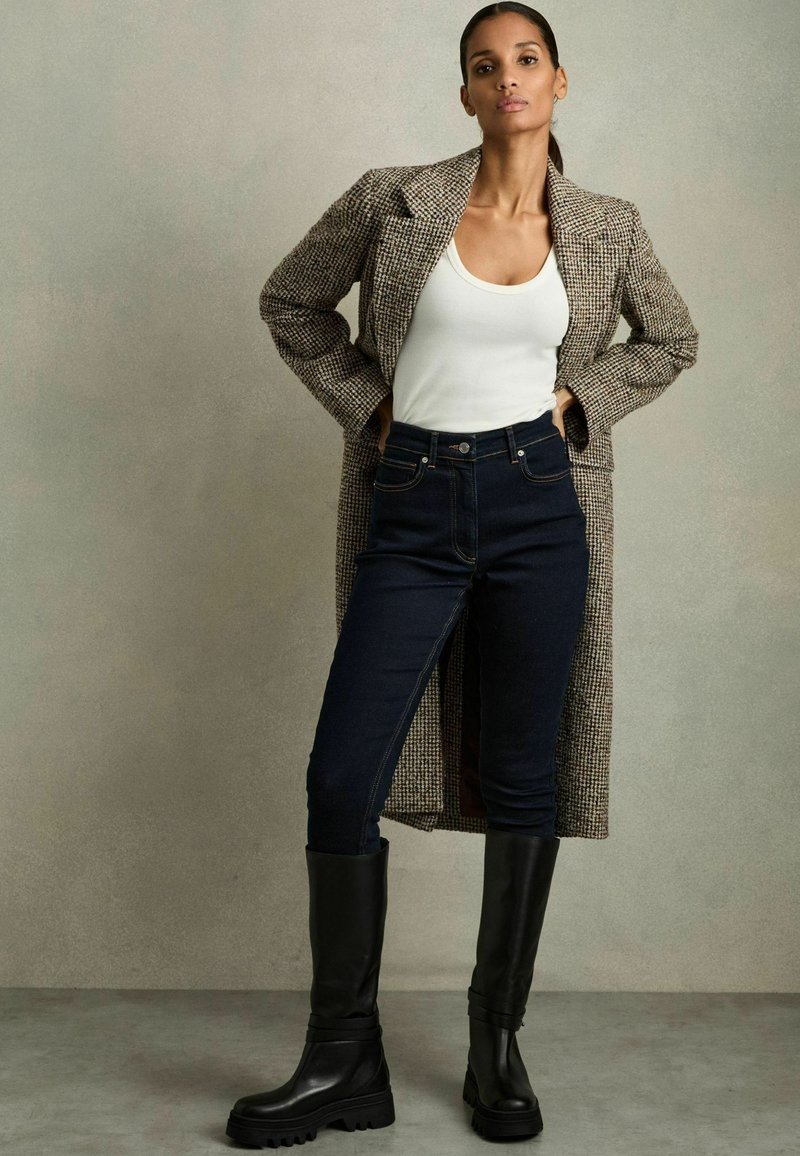 Brun rutigt långkappa, vit linne, mörkblå jeans och knähöga svarta stövlar med kraftiga sulor. Texturen varierar mellan materialen.